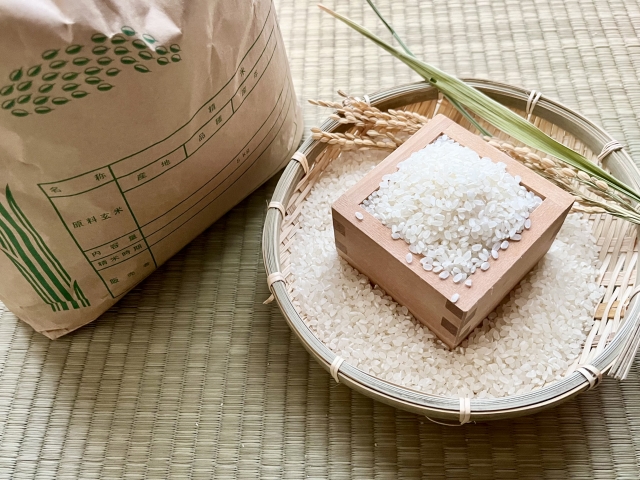 Japanese white rice displayed in a wooden masu on a bamboo tray beside a traditional rice bag and harvested rice stalks.