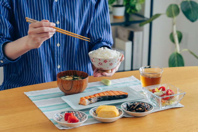 A person holding a bowl of white rice during a Japanese meal with grilled salmon, miso soup, tamagoyaki, pickles, and small side dishes.
