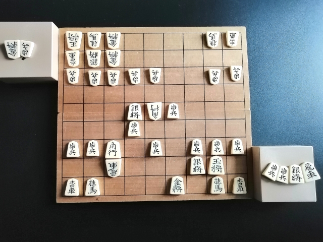 A strategic mid-game position in Shogi, showing pieces arranged on a wooden board with captured pieces on side tables.