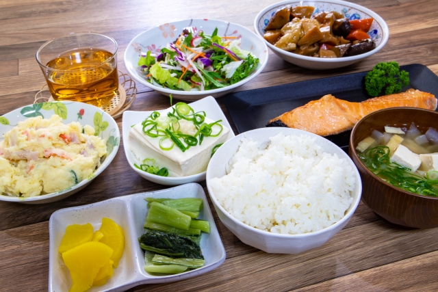 A Japanese meal with rice, miso soup, grilled salmon, tofu, pickles, salad, and small side dishes arranged on a table.