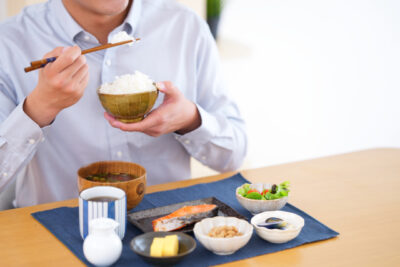 A Japanese meal with rice, miso soup, grilled fish, and small side dishes, with a person holding a rice bowl and eating with chopsticks