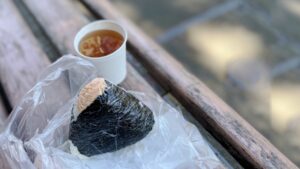 An onigiri rice ball on a bench beside a paper cup, showing rice as a simple meal outside the home.