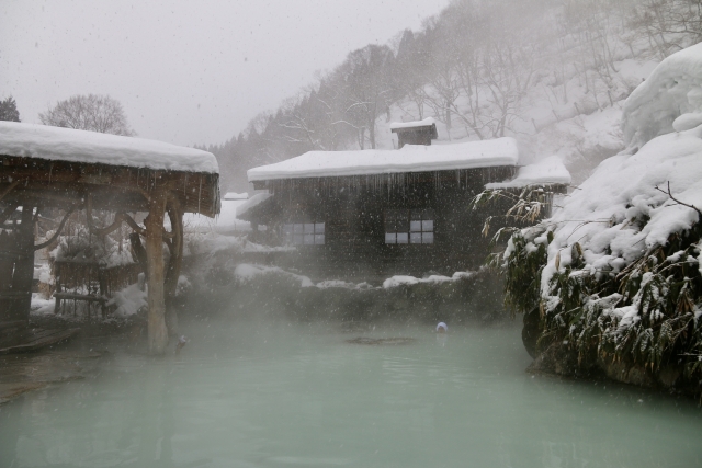 Snowy outdoor onsen bath with steam rising around traditional buildings in winter