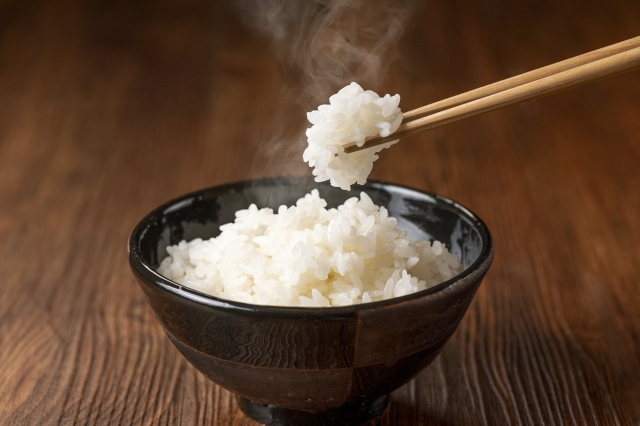 A bowl of freshly cooked Japanese short-grain rice with chopsticks lifting a small clump of steaming rice.