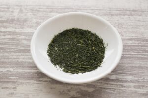 Deep-steamed sencha (fukamushi-cha) dry tea leaves in a white dish, showing fine broken leaf particles
