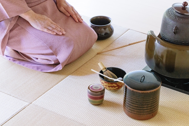Tea ceremony utensils on tatami next to an iron kettle, with the host seated in seiza
