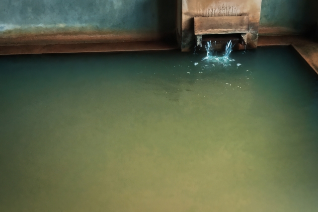 Indoor onsen bath with greenish mineral water and a stream of hot spring water flowing into the tub