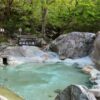 Outdoor onsen bath surrounded by rocks and forest, with steam rising from the water