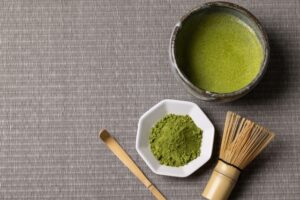 Matcha powder with a bamboo whisk and a bowl of prepared matcha.