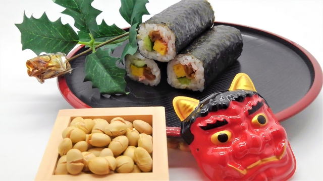 Setsubun items: roasted soybeans, ehomaki sushi rolls, an oni mask, and holly leaves with a sardine head
