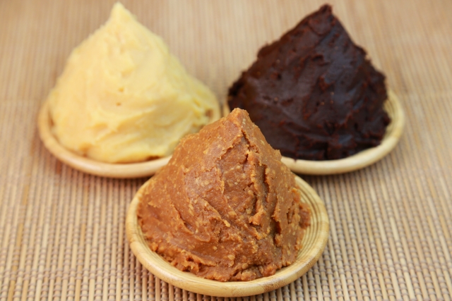Three varieties of Japanese miso paste in small wooden dishes, showing pale yellow, light brown, and dark reddish-brown colors.