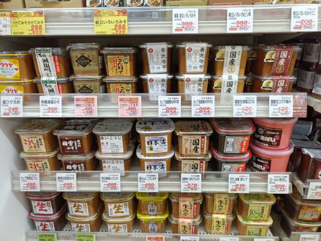 A Japanese supermarket shelf displaying many varieties of miso, including rice miso, additive-free miso, and reduced-sodium products.