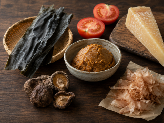 Umami-rich ingredients including kombu, dried shiitake mushrooms, miso, tomatoes, aged cheese, and katsuobushi arranged on a wooden table.