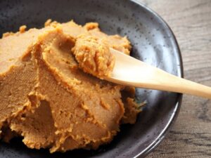 Reddish-brown Japanese blended miso in a dark ceramic bowl, with a wooden spoon holding a small portion.