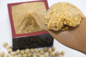 Light-colored Japanese rice miso on a wooden spoon, with soybeans in the background.