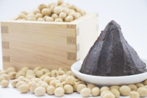 Dark brown Japanese soybean miso on a white dish, surrounded by whole soybeans and a wooden box.
