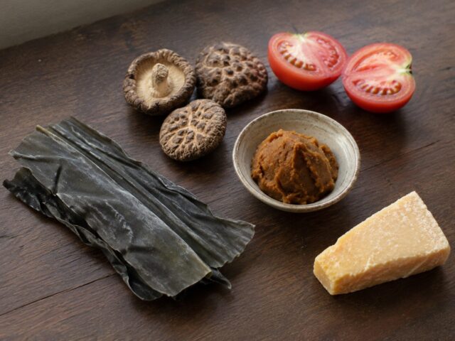 Kombu, dried shiitake mushrooms, tomatoes, miso, and aged cheese arranged on a wooden table as examples of umami-rich foods.