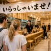 Foreign tourists entering a bright modern Japanese ramen shop as staff loudly greet them with “Irasshaimase!”