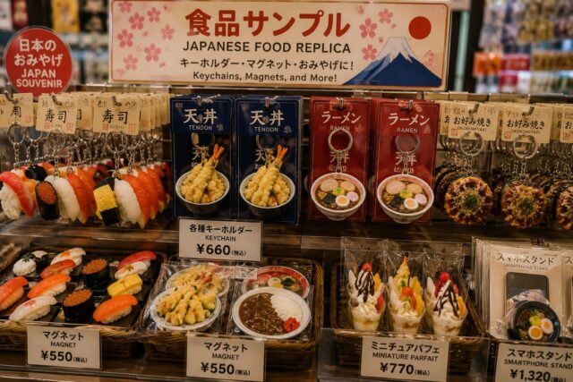 Japanese food replica souvenirs including sushi keychains, ramen magnets, miniature parfaits, and smartphone stands displayed in a gift shop