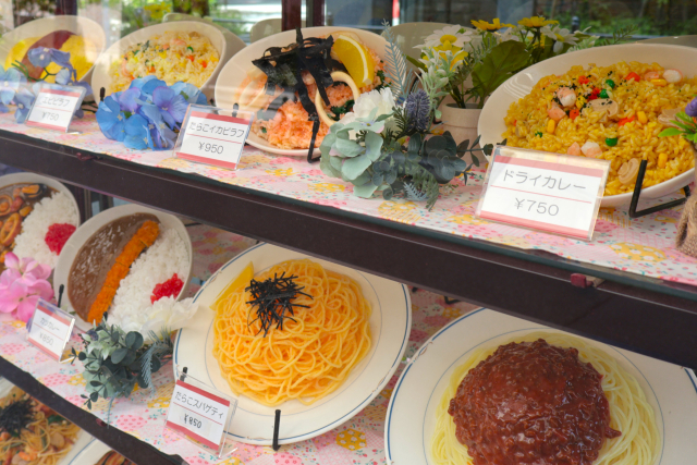 Foreign visitors looking at traditional Japanese restaurant food replicas displayed with menu labels and prices in a restaurant showcase