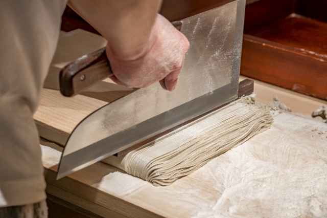 Soba craftsman cutting handmade buckwheat noodles with a traditional soba knife on a wooden workbench.