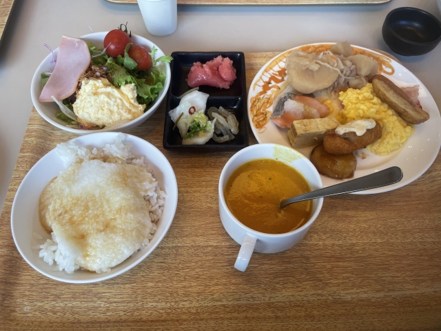 A hotel breakfast buffet tray combining Japanese and Western foods, including rice with grated yam, pickles, salad, eggs, sausage, and soup.