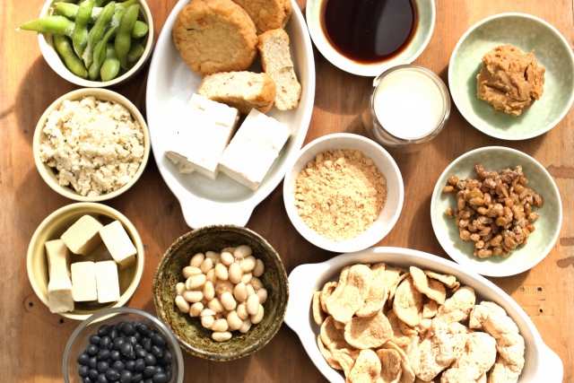 Japanese soybean foods including tofu, natto, miso, kinako, okara, soy milk, edamame, and whole soybeans arranged on a wooden table.