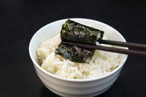 A small piece of seasoned nori held with chopsticks over a bowl of rice, showing how ajitsuke nori is often eaten with rice in Japan.