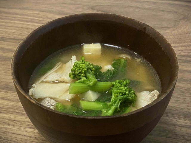 A bowl of miso soup with asparagus, greens, tofu, and other vegetables in a wooden bowl.