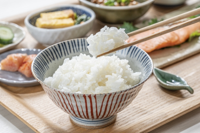 A bowl of steamed white rice in the foreground, with chopsticks lifting a bite and several Japanese side dishes arranged behind it. Rice is not counted as one of the three dishes because it forms the base of the meal. This can seem unusual to readers from cultures where the largest meat or fish dish is treated as the center. In many Japanese meals, rice provides the stable foundation around which everything else is arranged. It is mild, soft, and filling. The other dishes may be salty, savory, sour, sweet, or strongly seasoned. Rice balances those flavors and gives them a place within the meal. A piece of grilled fish may be quite salty on its own. Pickles may be too intense to eat in large amounts. Simmered vegetables may carry soy sauce, dashi, or sweetness. With rice, those foods become easier to combine. This is one reason the side dishes can remain small. They do not need to provide the whole meal by themselves.
