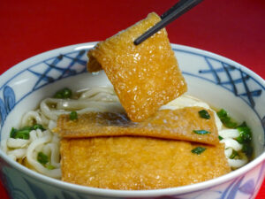 A piece of aburaage, or thin fried tofu, being lifted from a bowl of Japanese udon noodle soup.