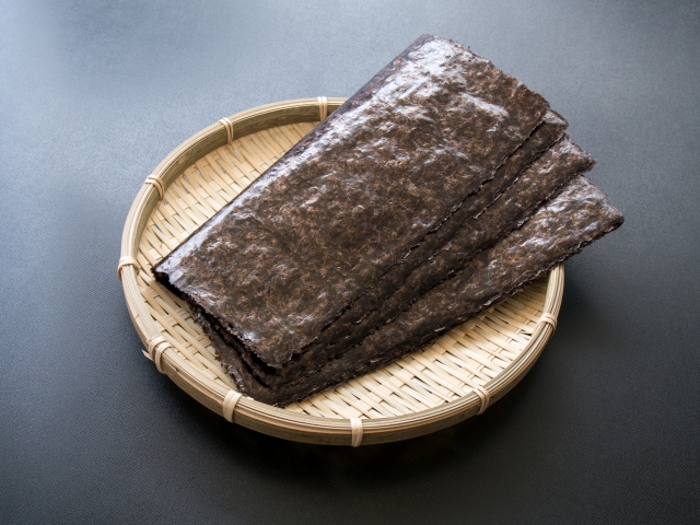 A stack of sheet nori on a bamboo tray, showing the dark color, texture, and uniform shape of dried seaweed sheets.
