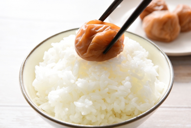 An umeboshi being placed on a bowl of white rice, showing how strongly flavored Japanese pickles balance plain rice.