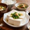 A Japanese home meal with hiyayakko tofu topped with green onion and ginger, served alongside rice, miso soup, soy sauce, and small side dish