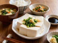 A Japanese home meal with hiyayakko tofu topped with green onion and ginger, served alongside rice, miso soup, soy sauce, and small side dish