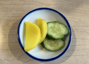 A small bowl of Japanese pickles, including takuan and cucumber, served in a modest portion with breakfast.