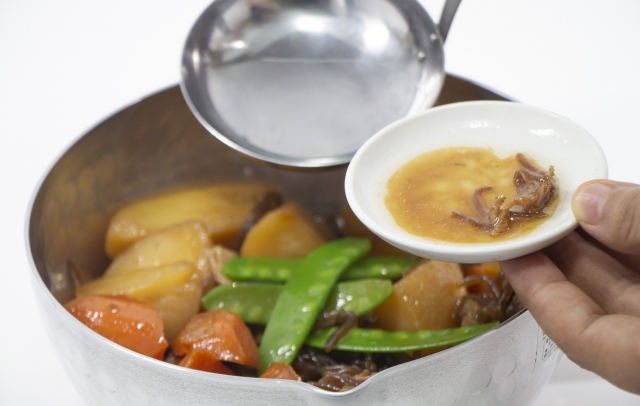 A small dish of cooking broth being held beside a pot of simmered vegetables and meat.