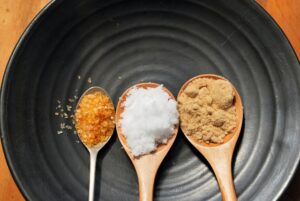 Three different sugars with distinct crystal sizes and textures displayed on spoons over a black plate