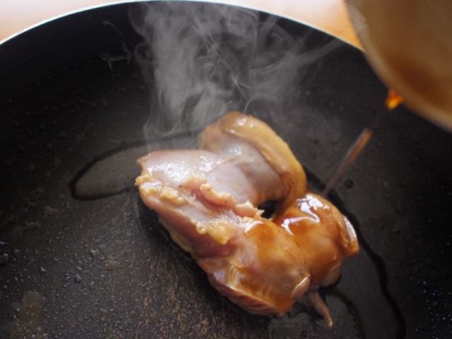 Seasoning being poured over chicken in a hot frying pan as steam rises during cooking.