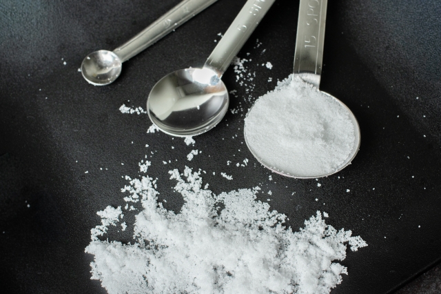 Metal measuring spoons with white sugar on a dark surface, illustrating volume measurements used in recipes